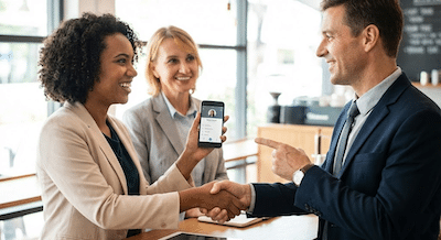 A candid photograph of two smiling business professionals, a woman and a man, shaking hands warmly in a brightly lit cafe. One person is holding up a smartphone displaying a contact card on the screen, while the other is pointing at the phone with a pleased expression. A third person in the background is looking at them with a smile. The scene conveys a quick, immediate exchange of business information.