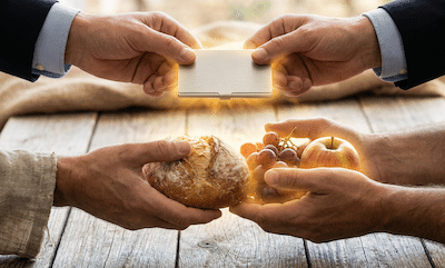 A close-up photograph focused on four hands. Two hands are exchanging a sleek, modern metal business card holder. Simultaneously, underneath the business cards, the other two hands are exchanging a rustic loaf of bread and fruit. Both the modern business items and the ancient food items are glowing with the same warm, golden energy, symbolizing that they are the same fundamental transaction of survival.