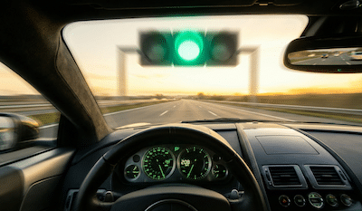 A dynamic, motion-blurred photograph taken from the perspective of a driver inside the cockpit of a high-performance luxury sports car. The dashboard gauges are illuminated with bright green light, showing the speedometer needle near maximum. Through the windshield, a massive, stylized traffic light hovering over an eight-lane empty highway shows a brilliant, glowing green light. The road ahead is wide open and stretches into a golden horizon. In the rearview mirror, other generic cars are visible but are small and heavily blurred, indicating they are being left far behind. The feeling is one of intense speed and unstoppable momentum.