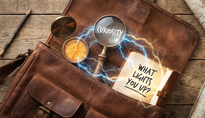 A flat-lay photograph on a rustic wooden table surface. An open, adventurer-style leather satchel reveals glowing conversational tools: an old-fashioned compass labeled "PASSION," a magnifying glass labeled "CURIOSITY," and a small notebook with the handwritten words "WHAT LIGHTS YOU UP?" in bold ink. Small electrical blue and gold sparks are jumping between the items in the bag.