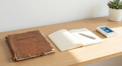 A minimalist, clean concept photograph representing a fresh start. On a clean white oak desk surface lies an old, worn-out business plan folder next to a brand new, open blank journal with a high-quality pen resting on the first page. Beside them is a sleek, modern prototype device suggesting innovation. A strong beam of natural sunlight hits the blank page. The mood is optimistic, clean, and ready for action.