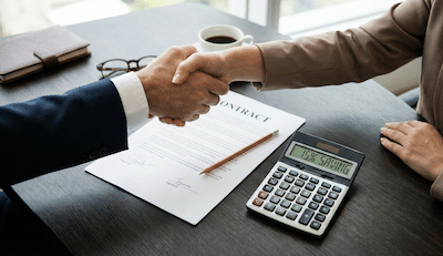 An overhead desk shot of a serious business negotiation being finalised. Two hands (one male, one female in business attire) are firmly shaking hands across a wooden table over a signed contract document. Resting on the contract is an exceptionally sharp pencil and a modern calculator displaying a green percentage "SAVING." The aesthetic is professional, focused, and decisive.