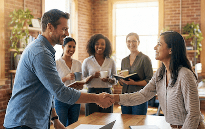A warm, candid photograph of a small business team (the "Clan") gathered for a meeting in a modern, sunlit office. The focus is on a genuine, smiling handshake between a business owner and a key employee, conveying trust and value. Other diverse team members are in the background, looking engaged and supportive. The atmosphere is loyal and positive.